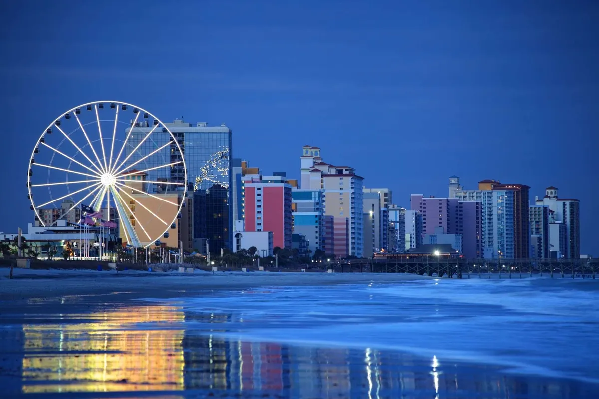 A vibrant view of the beachfront boardwalk, featuring a prominent Ferris wheel, colorful buildings, and shimmering reflections on the water. Families and visitors are seen strolling along the beach, enjoying amusement park rides, and dining at nearby restaurants, all under the warm glow of sunset.
