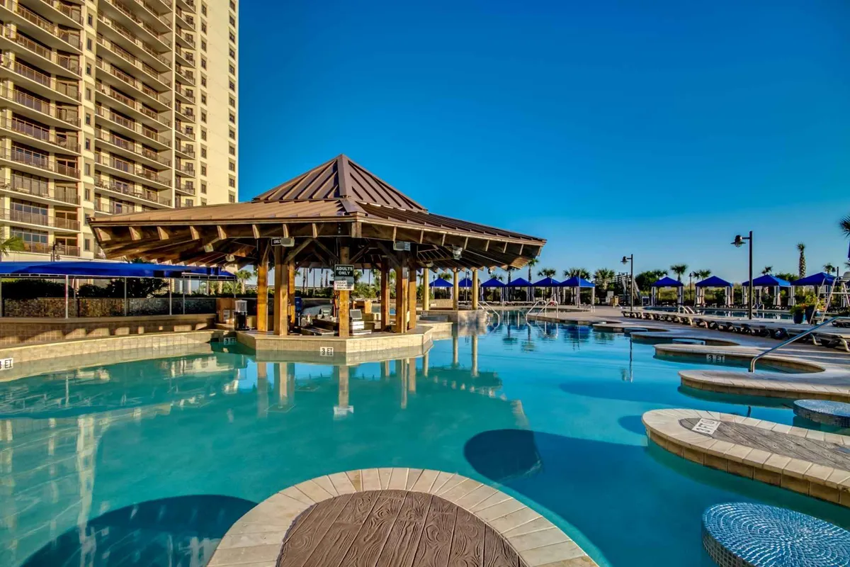 A vibrant resort pool area featuring a large swimming pool surrounded by lounge chairs, blue umbrellas, and a tiki-style gazebo. The mood is casual and enjoyable, suggesting a vacation-friendly environment with sunset views and opportunities for poolside cocktails and games.