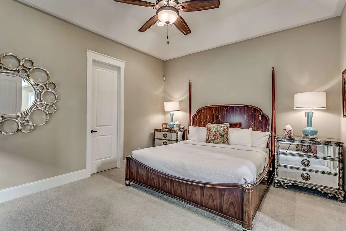 A calm and restful bedroom featuring a plush bed with decorative pillows, a mirrored nightstand, ceiling fan, and a wall mirror. The neutral tones and soft lighting create a cozy, inviting atmosphere perfect for relaxation and unwinding after a long day.