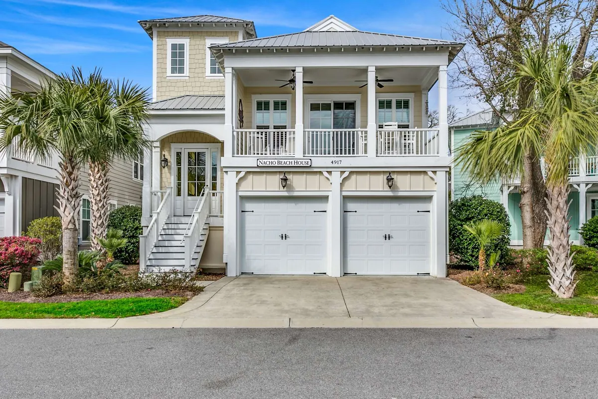 The image showcases the inviting exterior of a spacious single-family home in North Myrtle Beach, featuring a charming front porch with white railings, a welcoming staircase, swaying palm trees, and large garage doors. The scene is bathed in warm sunset light, hinting at relaxing evenings and vibrant gatherings. The porch offers space for family or friends to gather, sip morning coffee, or enjoy cheerful outdoor barbecues.