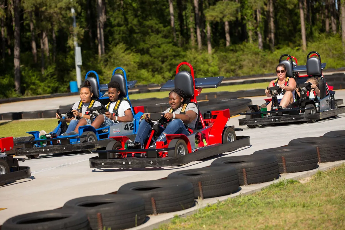A dynamic outdoor scene showing participants racing go-karts around a twisting track, surrounded by lush trees. The action is lively, with families cheering and celebrating, capturing the excitement of a day spent at a popular local attraction.