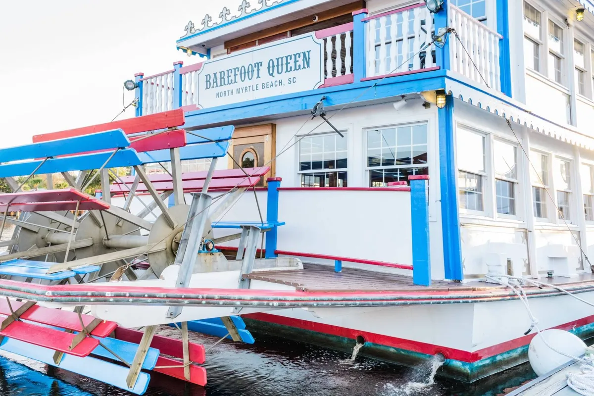 A classic paddle-wheel riverboat, the 'Barefoot Queen,' with colorful exterior architecture and clear signage, is docked at the waterfront. Guests are seen enjoying dining and sightseeing cruises, with the mood relaxed and cheerful, perfect for group gatherings or themed events.
