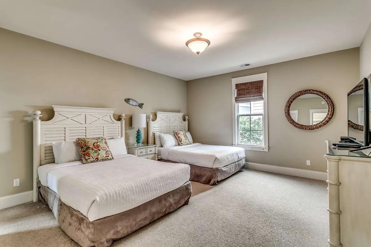 A cozy bedroom with two beds, each adorned with decorative pillows. A large mirror, window with blinds, and a spacious chest of drawers enhance functionality and comfort. Gentle morning sunlight spills in, creating a warm and inviting setting for conversation or relaxation.