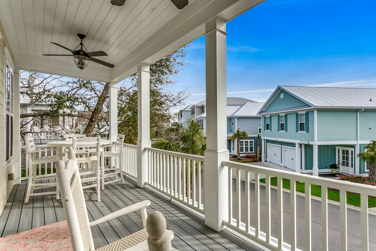 A spacious covered balcony furnished with comfortable porch seating overlooks a tranquil neighborhood setting. The space is enhanced by a ceiling fan for comfort, and the scene is bathed in warm natural light, with glimpses of trees and a soft evening or morning ambiance.