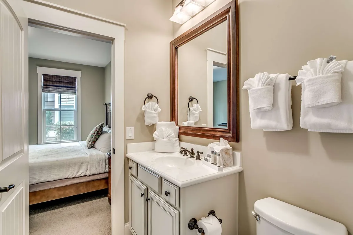 A bright bathroom featuring a vanity with sink, a large mirror, soft towels on a rack, and a window streaming in natural light. A glimpse of a bed through the doorway connects the bathroom to the adjacent bedroom, underscoring the suite-like layout.