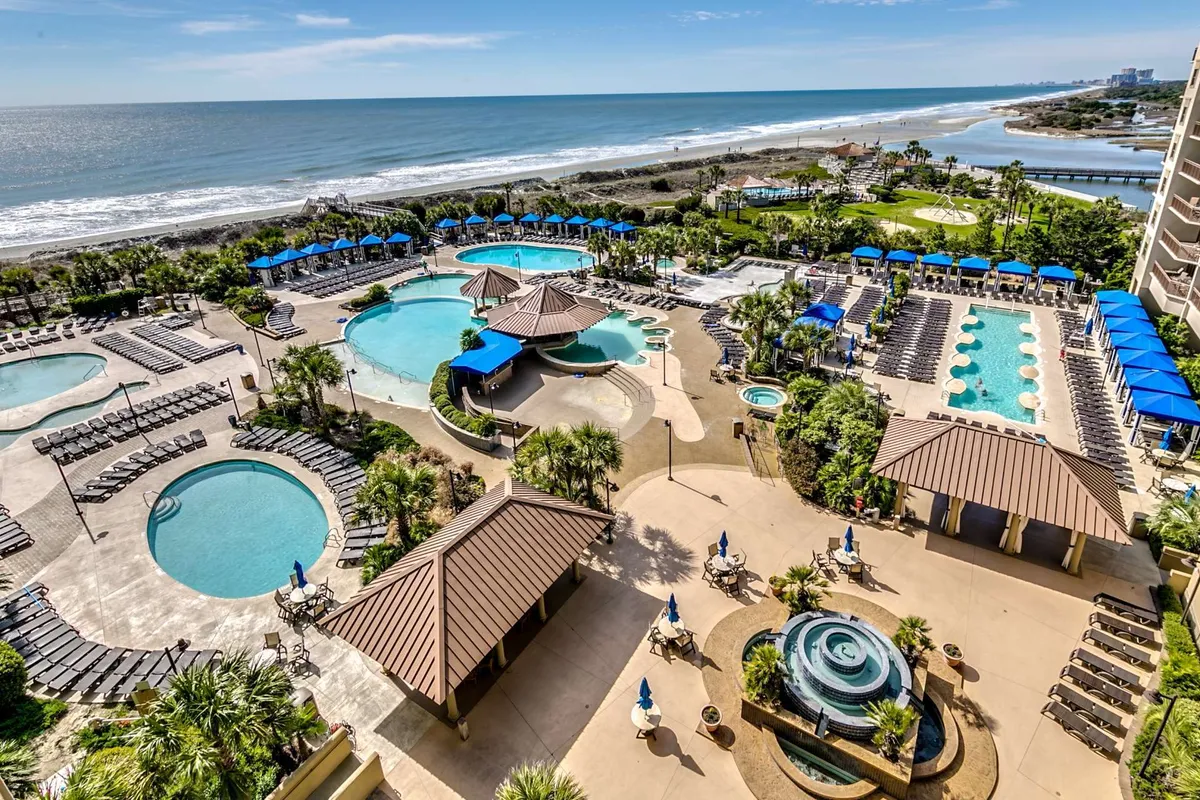 An expansive outdoor pool complex with multiple swimming pools, palm trees, beach access, comfortable seating areas, and cabanas. The scene is serene and vacation-friendly, hinting at poolside relaxation and social gatherings under the sun or stars.
